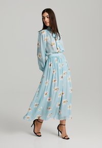 Robe florale bleu clair avec des manches longues et un col à nouer. La jupe présente une silhouette fluide, ornée de fleurs blanches et d'une texture de tissu douce.
