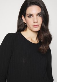 Top preto de manga longa com textura estriada e decote redondo, apresentando uma textura suave e um brilho sutil. Acessorizado com brincos de argola dourados.