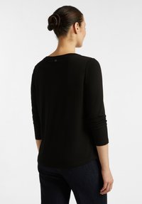 Černé tričko s dlouhými rukávy a kulatým výstřihem, vyrobené z měkké tkaniny a s přiléhavým střihem. Na zadním výstřihu se nachází malé logo.