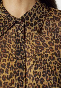 Camicia con stampa leopardata caratterizzata da un colletto, chiusura frontale con bottoni e taschino sul petto. Il tessuto è semi-trasparente con diverse tonalità di marrone e nero.