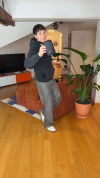 Melna auduma plecu soma ar priekšējo kabatu, ko tur cilvēks pelēkā kapučjakā un gaiši pelēkos biksēs, stāvot telpā ar augu puķuzirņiem.