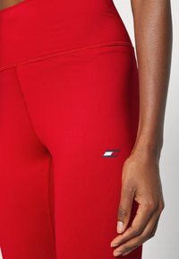 Leggings rossi realizzati in materiale elasticizzato con cuciture visibili. Presentano un piccolo logo sul lato con una combinazione di colori blu, bianco e rosso.