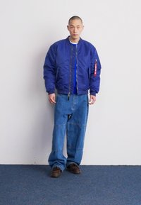 Chaqueta bomber azul con cierre de cremallera y bolsillos laterales, combinada con pantalones de mezclilla azules de gran tamaño y zapatos marrones.