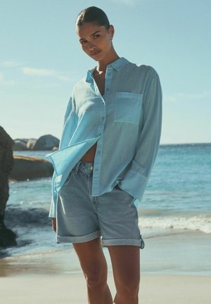 Light blue, button-up shirt with a chest pocket, paired with rolled-up denim shorts. Beach setting with water and rocks in the background.