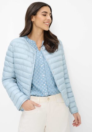 Frau lächelnd, trägt eine hellblaue gesteppte Jacke über einer gemusterten blauen Bluse und beige Hosen, steht vor weißem Hintergrund.