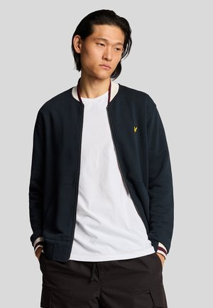 Marine kleurige zip-up jas van zacht materiaal, met ribgebreide manchetten met kastanjebruine en witte strepen, en een klein geel logo op de borst.