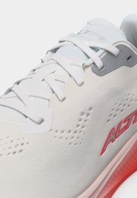 Λευκό αθλητικό παπούτσι με λευκά κορδόνια, γκρι λεπτομέρειες, διάτρητο επάνω μέρος, κόκκινο λογότυπο "ALTRA" και εμφανές κοκκινωπό υφασμένο άκρο σόλας.