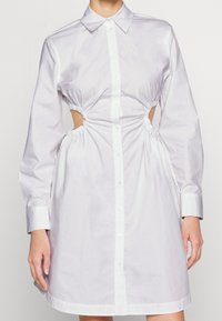 Vit bomullsklänning med knäppning framtill, långa ärmar och insydda sidoutskärningar. Har en klassisk krage och knä-lång fåll.