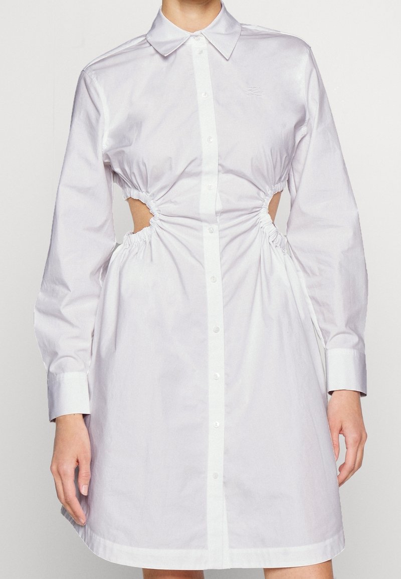 Vit bomullsklänning med knäppning framtill, långa ärmar och insydda sidoutskärningar. Har en klassisk krage och knä-lång fåll.