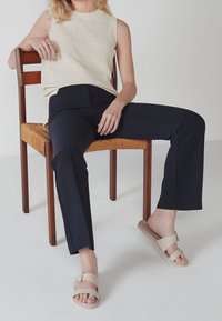 Beige geribbelde mouwloze top, marineblauwe slim-fit broek en beige sandalen. Zittend op een houten stoel met een geweven zitting. Eenvoudig, modern ontwerp.