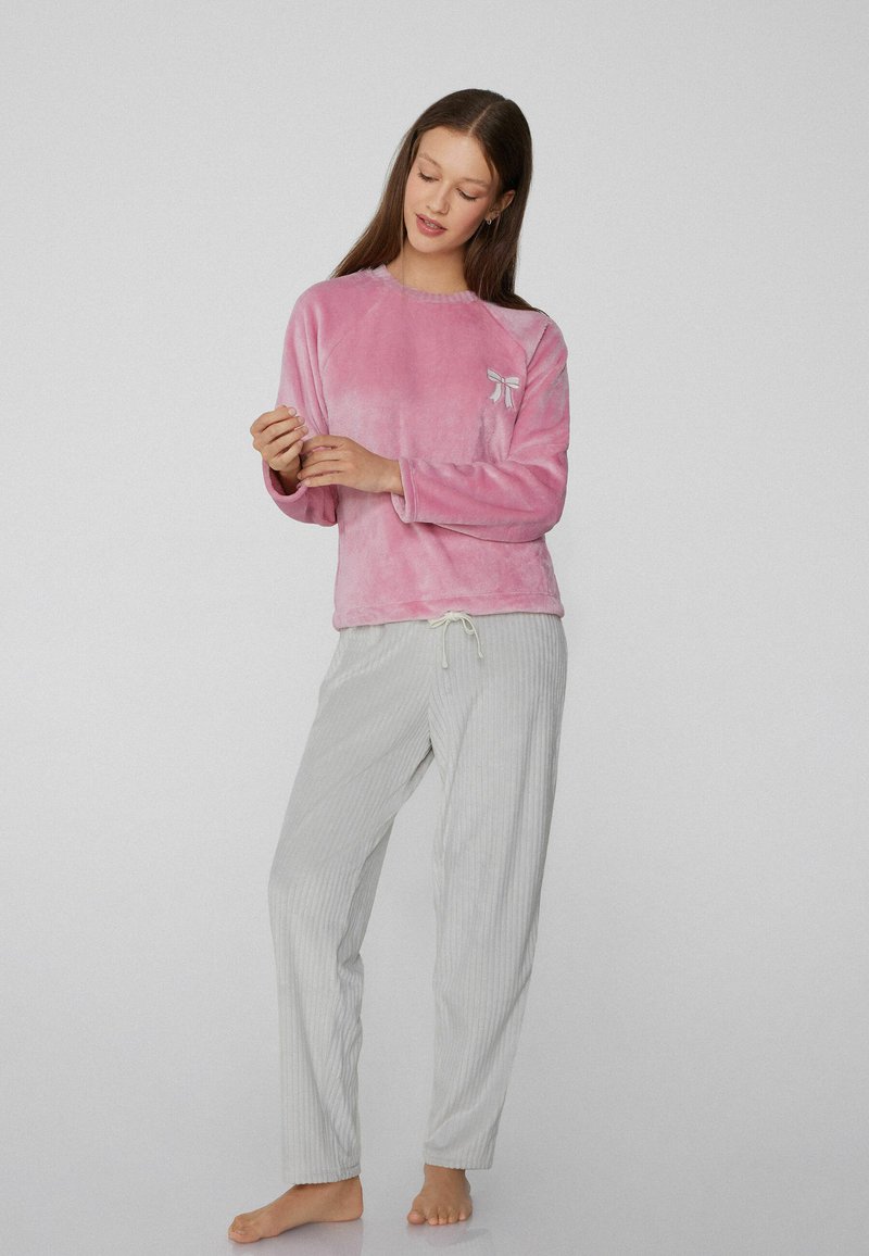 Camiseta rosa de felpa de manga larga con un pequeño lazo decorativo, combinada con pantalones a rayas gris claro con cinturilla elástica y cordón.