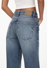 Světle modré široké džíny s vysokým pasem, se zadními kapsami a hnědým koženým štítkem na pasu. Textura denimu je viditelná.