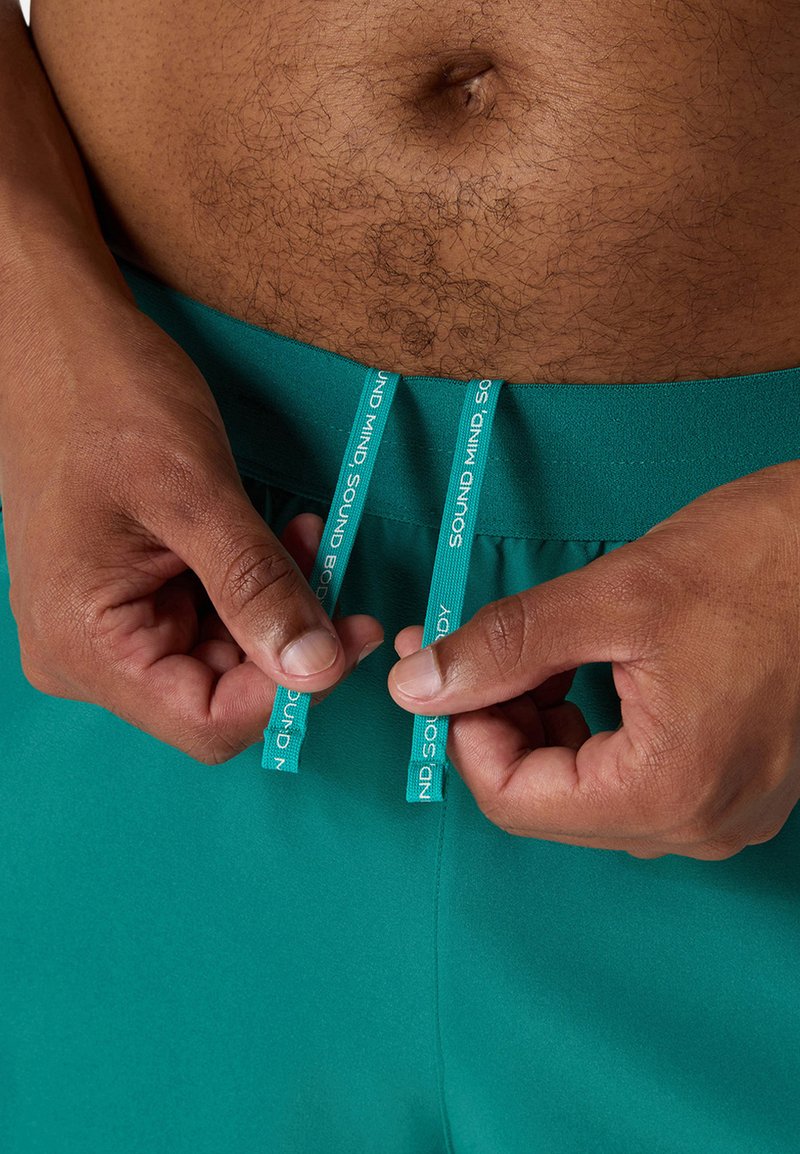 Teal sportshorts met een gladde textuur, voorzien van een trekkoord met het label "SOUND MIND SOUND BODY." Handen grijpen de trekkoorden.