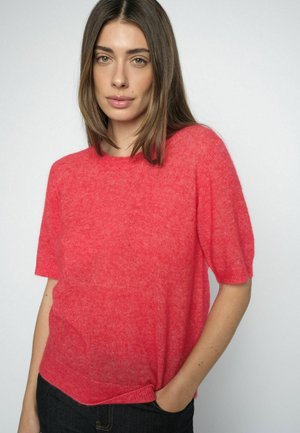 Frau mit glatten braunen Haaren, die einen roten Kurzarmpullover und schwarze Hosen trägt, mit einer Hand in der Tasche vor schlichtem Hintergrund.