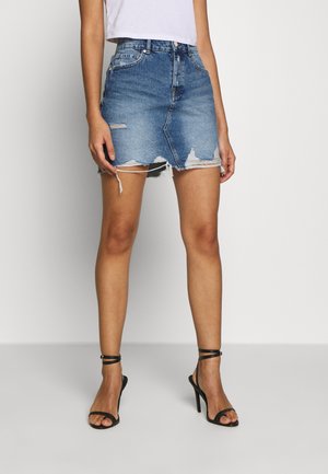 Kvinde iført en slidt blå denim mininederdel, hvid croppet top og sorte højhælede sandaler med stropper, stående foran en ensfarvet baggrund.