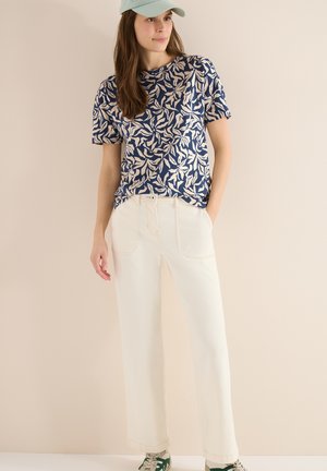 Jeune femme portant une chemise à motifs de feuilles bleu et beige, un pantalon large crème, des baskets vertes et blanches, et une casquette vert clair.