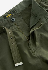 Pantaloni cargo de culoare verde măsliniu, dintr-un material texturat, cu fermoar metalic, închidere cu buton și curea din material. Eticheta indică marca și mărimea 32.