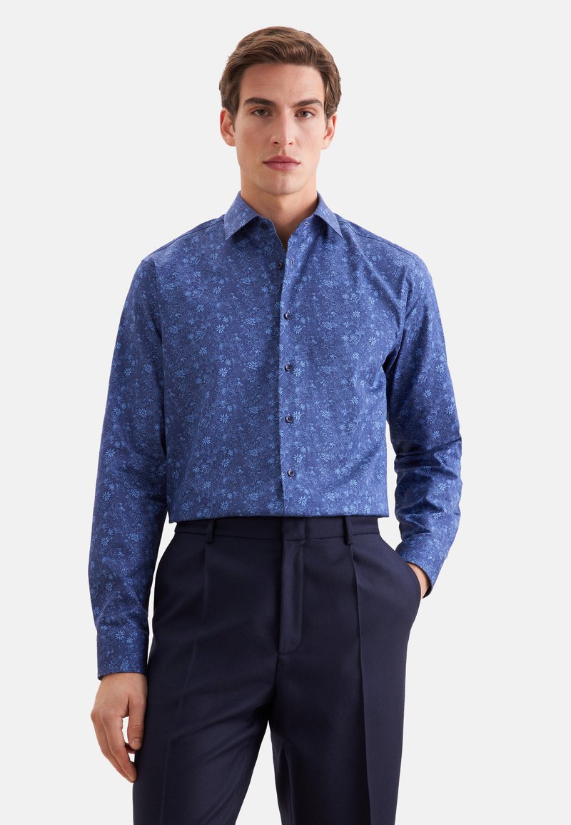 Homme portant une chemise bleue à manches longues à motif floral avec un pantalon sombre, debout avec une main dans la poche sur fond blanc.