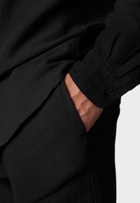 Schwarzes Shirt aus weichem Stoff mit subtiler Textur, ausgestattet mit einem knöpfbaren Ärmel und einer Seitentasche mit gebogener Öffnung.