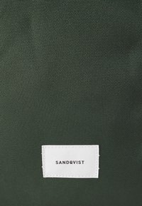 Sac en tissu vert foncé avec une finition texturée. Il présente une étiquette blanche avec "SANDQVIST" cousu en noir, positionnée en bas.