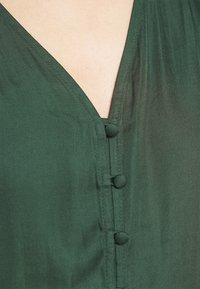 Blusa verde scuro con scollo a V, leggero lucido e bottoni rivestiti in tessuto lungo la parte frontale per la chiusura. Tessuto liscio.