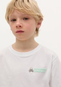 T-shirt bianco in cotone, con logo verde menta ricamato nella zona sinistra del petto, raffigurante teschio e ossa incrociate. Design a girocollo.