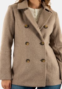 Cappotto doppiopetto in misto lana beige con grandi bottoni scuri, colletto a rever e texture liscia. Dotato di tasche laterali e vestibilità su misura.
