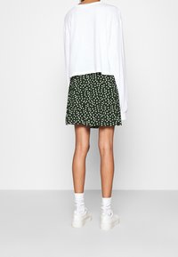 Haut blanc à manches longues associé à une jupe verte à pois blancs ; le mannequin porte des baskets blanches et des chaussettes, jambes nues apparentes.