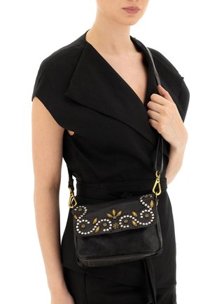 Donna con top nero senza maniche tiene una piccola borsa a tracolla nera decorata con borchie dorate e argento a motivi floreali.