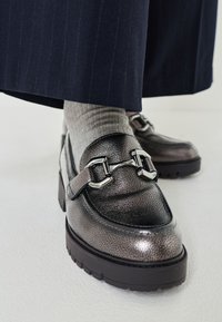 Metallic leather loafers feature a textured surface, silver hardware detail, and a thick black sole. Worn with grey ribbed socks and navy pants.