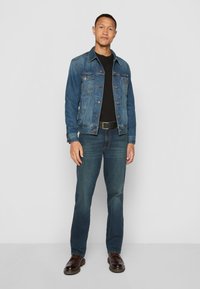 Denim jacket in medium blue with button closures, worn over a black shirt. Dark blue jeans and brown leather shoes complete the outfit.