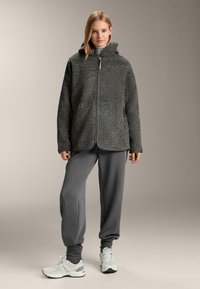 Siva flis jopica z zadrgo, kapuco in stranskimi žepi, kombinirana s sivimi trenirkami in belimi supergami. Gladek tekstura, sproščen kroj.