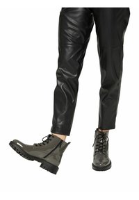 Personne portant un pantalon en cuir noir et des bottines gris brillant à lacets avec fermetures éclair latérales, un pied légèrement levé.