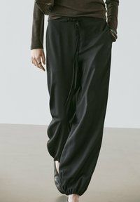 Kvinde iført løstsiddende sorte bukser med elastiske manchetter, en brun langærmet bluse, sorte sko og armbånd på det ene håndled.