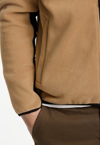 Chaqueta de forro polar con cremallera en color marrón claro con ribetes negros, cuenta con bolsillos laterales y un acabado texturizado, llevada sobre pantalones marrones estampados.