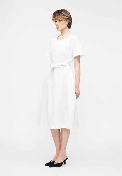 Γυναίκα που στέκεται πλαγίως, φορώντας λευκό φόρεμα έως το μέσο της γάμπας με κοντά μανίκια και ζώνη στη μέση, καθώς και μαύρα μυτερά παπούτσια με τακούνι.