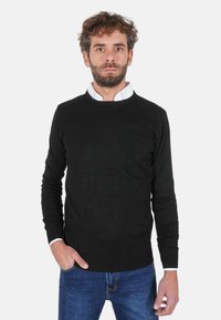 Maglione nero a girocollo realizzato in tessuto liscio, abbinato a una camicia bianca con colletto sotto e jeans di denim blu. Vestibilità casual.