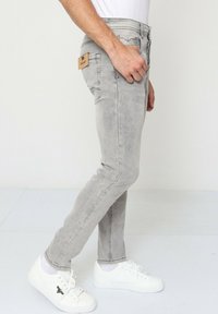Jean en denim gris présentant une coupe slim, un design à cinq poches et une étiquette en cuir à l'arrière. Portés avec des baskets blanches.