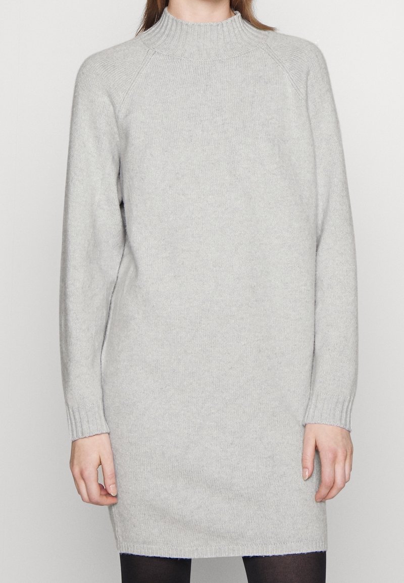 Femme portant une robe-pull en maille gris clair unie à manches longues avec un col montant côtelé et des leggings noirs.