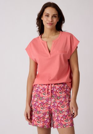 Femme portant un haut corail rose à manches courtes et un short à motifs floraux rose, vert et blanc, debout devant un fond uni.