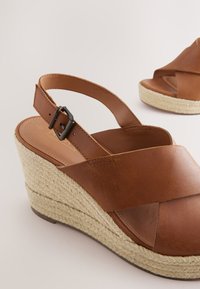 Sandales compensées en cuir marron avec un design à bretelles croisées, dotées d'un talon en jute tissé et d'une bride de cheville ajustable avec une boucle métallique.