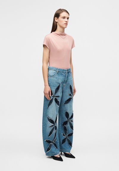 Haut ajusté rose clair à col montant, assorti à un jean denim bleu à jambes larges avec broderies florales noires et bords effilochés.