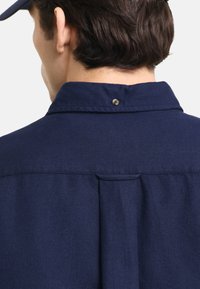 Camicia color blu navy con colletto, caratterizzata da una trama a spina di pesce, dettaglio con piega sul retro e chiusura a scatto sul colletto per un ulteriore interesse di design.