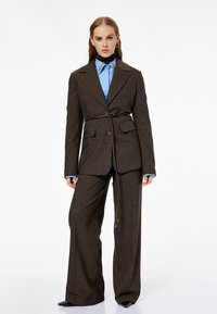 Brun ternet blazer med bælte, parret med en blå skjorte med knapper. Har brede brune bukser og blanke sorte sko.