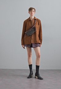 Veste oversize en daim marron portée sur une robe en tissu à motifs, associée à des bottes noires à talons hauts jusqu'aux genoux et à un petit sac en bandoulière.