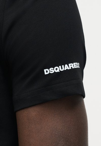 Μαύρο κοντομάνικο πουκάμισο σε σκουρόχρωμο δέρμα με λευκή επιγραφή "DSQUARED2" τυπωμένη κοντά στο τελείωμα του μανικιού.
