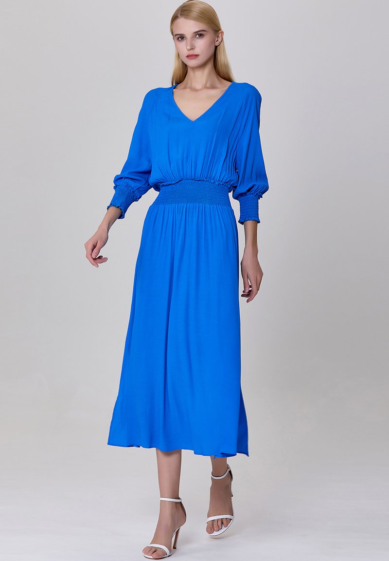 Robe maxi bleue avec un décolleté en V, des manches bouffantes et une taille smockée. Tissu lisse avec une silhouette fluide, accompagnée de talons à bride blanche.