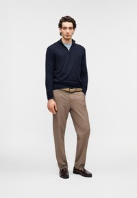 Pull en tricot bleu marine à fermeture éclair porté par-dessus une chemise bleu clair, associé à un pantalon beige clair et des mocassins marron. La tenue est bien ajustée.