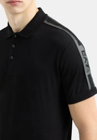 Polo shirt nera con maniche corte, caratterizzata da un accento testurizzato nero e grigio sulle spalle e cinque bottoni frontali.