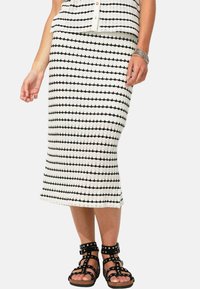 Jupe tricotée en blanc avec des rayures horizontales noires, présentant une coupe ajustée. Associée à des sandales noires à lanières avec des accents cloutés.